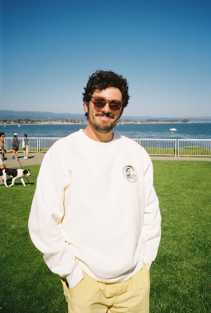 Sam Isenhart, smiling outdoors in a white sweatshirt and sunglasses with water and hills behind.